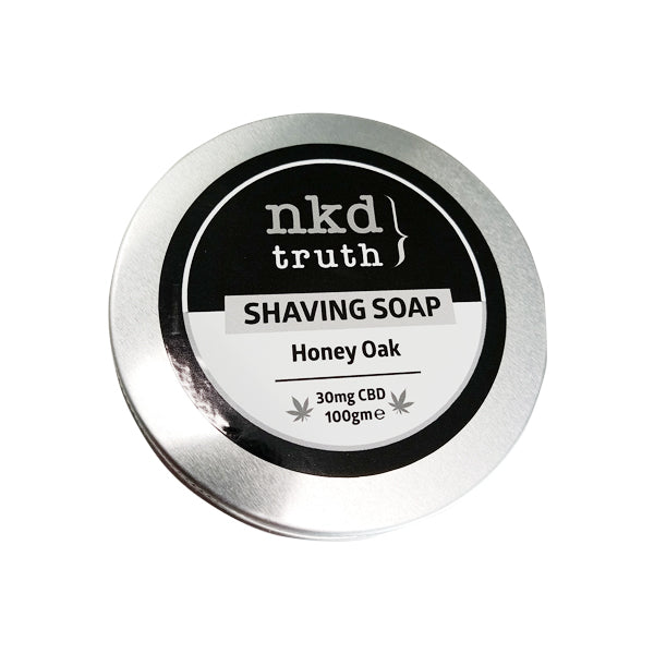 A sleek silver round metal tin container featuring an elegant black and white label reads 'NKD 30mg CBD Speciality Shaving Soap 100g - Honey Oak.' Highlighting its luxurious Honey Oak fragrance, the premium quality ingredients include 30mg of CBD and a net weight of 100 grams.